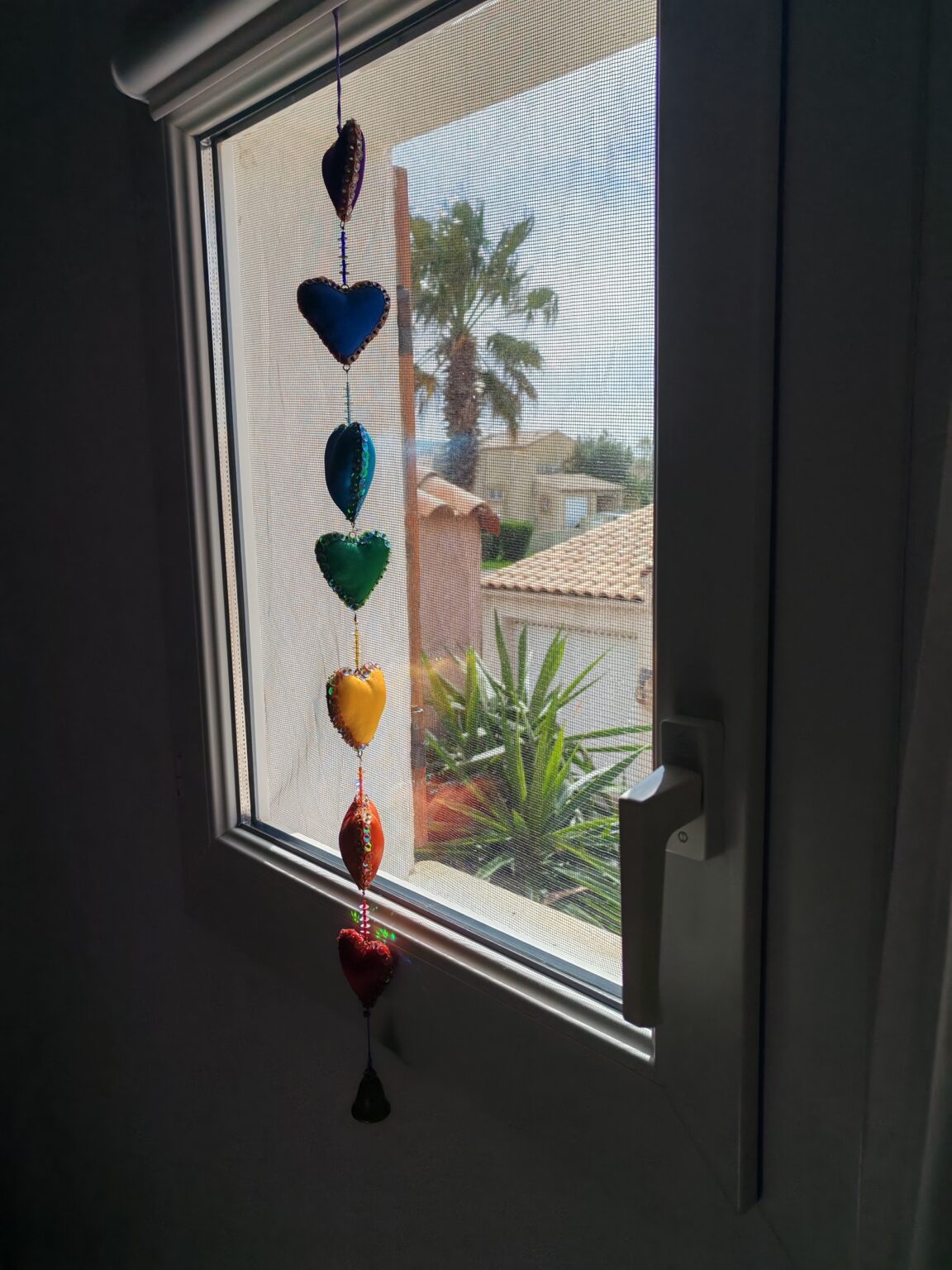 Bunte Herzanhänger in einem Fenster, draußen eine Palme